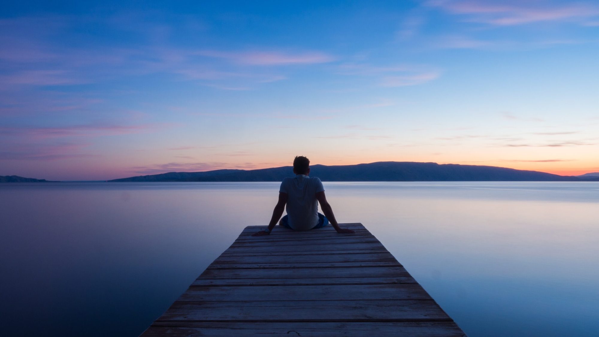 A person sitting alone at the end of a wooden jetty, looking out over still water